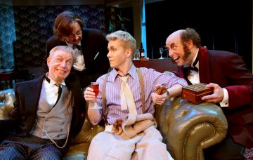 Neljä vintage-asuihin pukeutunutta näyttelijää istuu vihreällä sohvalla. Yksi pitää kädessään juomaa, toinen esittelee puulaatikkoa, ja muut hymyilevät ja nojaavat sisäänpäin, mikä viittaa vilkkaaseen, humoristiseen keskusteluun teatteriympäristössä.