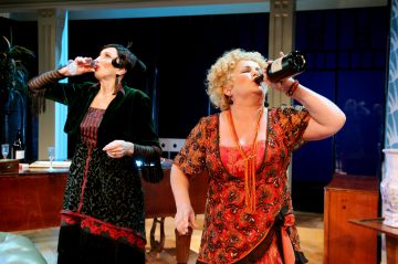 Kaksi värikkäisiin vintage-mekkoihin pukeutunutta naista seisoo olohuoneessa ja juo kumpikin viinipullosta. Kohtaus vaikuttaa vilkkaalta, mikä viittaa teatteriesitykseen tai komedialliseen hetkeen.