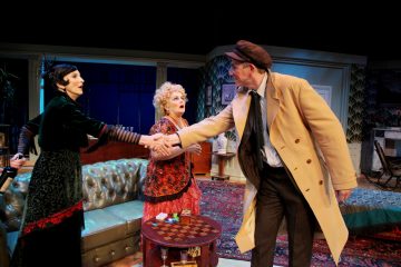 Lavalla on kolme vintage-asuihin pukeutunutta näyttelijää; vihreäpukuinen nainen ja rusketustakkinen mies kättelevät toisiaan, kun taas toinen punapukuinen nainen katselee hämmästyneenä. Lavasteena on klassinen olohuone, jossa on sohva ja kuviollinen matto.