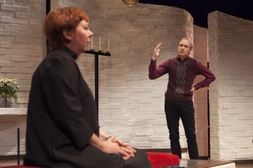 Lyhyet punaiset hiukset omaava henkilö istuu vasempaan päin, kun taas toinen henkilö seisoo hänen takanaan ja elehtii yhdellä kädellä. He ovat lavasteessa, jossa on valkoiset tiiliseinät ja minimaaliset koristeet.