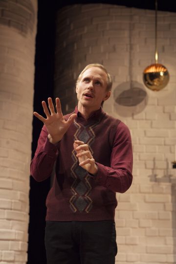 Mies, jolla on ruskeankeltainen villaliivi ja viininpunainen paita, elehtii nostamalla toisen kätensä ylös ja puhuu kiihkeästi kaarevien valkoisten tiiliseinien ja roikkuvan kultaisen pallon muodostaman taustan edessä.