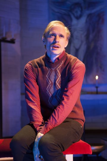 Vaaleat hiukset ja viikset omaava mies, jolla on punainen paita ja kuviollinen villaliivi, istuu penkillä kädet ristissä. Sinisellä valaistulla taustalla näkyy syttynyt kynttilä ja epäselvä taideteos.