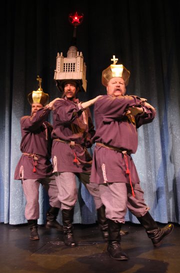 Lavalla esiintyy kolme keskiaikaiseen tyyliin pukeutunutta henkilöä, joilla kullakin on suuri, koristeellinen, rakennusten tai tornien muotoinen päähine, ja jotka seisovat rivissä kädet ristissä sinisen verhon edessä.