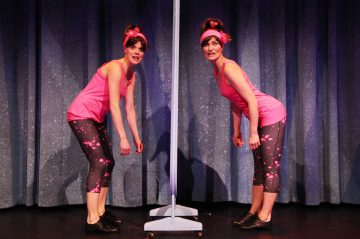 Kaksi naista, jotka ovat pukeutuneet samalla tavalla vaaleanpunaisiin toppiin, kuviollisiin legginsseihin ja vaaleanpunaisiin otsanauhoihin, seisovat pystysuoran peilin molemmin puolin ja jäljittelevät toistensa poseerausta näyttämöllä olevan sinisen verhon edessä.