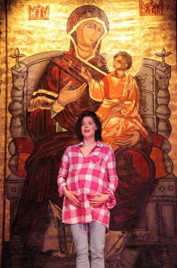 Vaaleanpunaiseen ruutupaitaan ja farkkuihin pukeutunut nainen seisoo raskaana olevaa vatsaansa pidellen suuren, kultaisen uskonnollisen Madonna ja lapsi -maalauksen edessä.