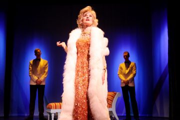 Lumoava nainen, jolla on oranssi, kimalteleva mekko ja valkoinen turkistukka, seisoo näyttämön keskellä dramaattisessa valaistuksessa, ja hänen takanaan on kaksi valkoisiin takkeihin ja mustiin housuihin pukeutunutta miestä. Taustalla näkyy valkoinen penkki, jossa on raidalliset tyynyt.