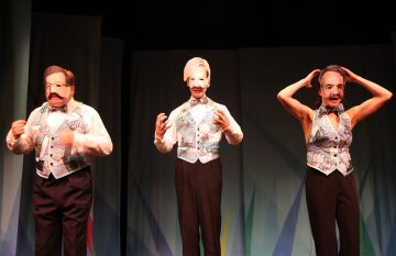 Lavalla seisoo kolme liiveihin ja rusetteihin pukeutunutta ihmistä, joilla kullakin on eri suuri naamio, joka kuvaa vakavia kasvoja. Taustaa koristavat abstraktit, värikkäät muodot.