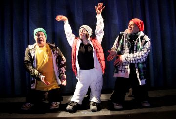 Kolme värikkäisiin talvivaatteisiin pukeutunutta ihmistä esiintyy lavalla tummansinisen verhon edessä. Keskellä olevalla henkilöllä on dramaattisesti kohotetut kädet, kun taas muut elehtivät käsillään ja ilmaisevat eloisia tunteita.