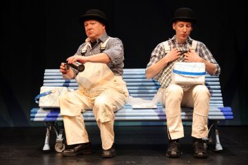 Kaksi keilahattuista ja haalarimiestä istuu sinisellä penkillä. Toisella on pullo ja lounasrasia kädessään, kun taas toinen säätää henkseleitään ja pitää lounasrasiaa. Molemmilla on vakavat ilmeet, ja heillä on samanlaiset maalitahrojen värittämät asusteet.