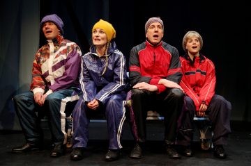 Neljä värikkäisiin talvitakkeihin, hattuihin ja urheiluvaatteisiin pukeutunutta ihmistä istuu vierekkäin näyttämön penkillä katsoen eteenpäin yllättynein tai lumoutunein ilmein. Tausta on tumma, mikä korostaa ryhmää.