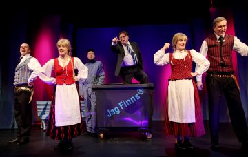 Lavalla kuusi näyttelijää, viisi värikkäissä perinteisissä skandinaavisissa asuissa ja yksi puvussa, poseeraavat energisesti mustan, Jag Finns® -merkinnällä varustetun roskiksen ympärillä, ja taustalla on dramaattinen violetti valaistus.