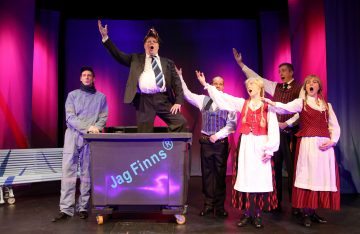 Kuusi ihmistä lavalla esiintyy teatterillisesti. Yksi mies puvussa ja kruunussa seisoo roskiksessa, jossa lukee Jag Finns. Muut, erilaisissa puvuissa, myös perinteisissä asuissa, elehtivät ja laulavat dramaattisesti värikkäiden näyttämövalojen alla.
