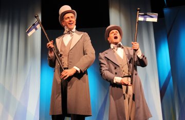 Kaksi harmaisiin pukuihin ja silinterihattuihin pukeutunutta miestä pitelee Suomen lippuja ja hymyilee esiintyessään lavalla, taustalla sinivalkoiset verhot.