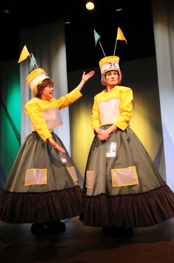 Kahdella lavalla olevalla henkilöllä on päällään omituiset puvut, joissa on keltaiset yläosat, suuret tummat hameet, selkeät taskut ja korkeat hatut, jotka on koristeltu numeroilla ja lipuilla. Toinen henkilö elehtii vilkkaasti toista kohti, joka seisoo kädet ristissä.