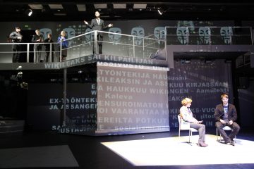 Teatterilava, jossa kaksi ihmistä istuu ja keskustelee valonheittimen alla, kun taas kolme ihmistä seisoo ylätasanteella. Taustalle ja lattiaan heijastetaan tekstiä ja kuvia, kuten Guy Fawkes -naamioita ja WikiLeaksiin liittyviä sanoja.