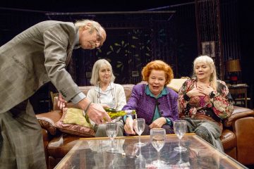 Vanhempi mies kaataa viiniä kahvipöydällä oleviin laseihin, kun kolme vanhempaa naista istuu sohvalla hymyillen ja lämpimästi reagoivina viihtyisässä, näyttämömaisessa olohuoneessa.