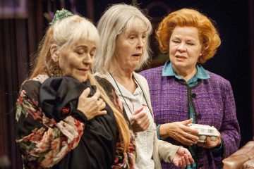 Kolme vanhempaa naista seisoo lähekkäin sisätiloissa. Yhdellä on kädessään mustaan kankaaseen kääritty nippu, toisella on valkoinen paita ja yllättynyt ilme, ja kolmas, punatukkainen, violettiin takkiin pukeutunut nainen pitää kädessään pientä valkoista astiaa.