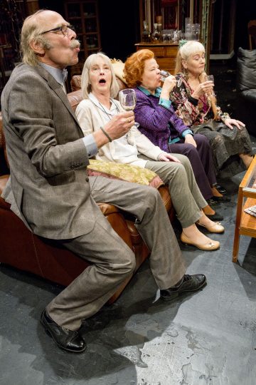 Neljä vanhempaa aikuista istuu sohvalla, jokaisella on lasi kädessään ja he reagoivat hämmästyneinä tai huvittuneina. Pukumies nojaa eteenpäin, ja hänen vieressään olevilla kolmella naisella on eloisa ilme. Ympäristö vaikuttaa viihtyisältä olohuoneelta.