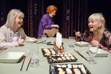 Kolme vanhempaa naista istuu pöydän ympärillä sushi-tarjottimien kanssa ja nauraa yhdessä. Yksi nainen seisoo, kun kaksi muuta istuvat, ja kaikki vaikuttavat iloisilta ja keskustelevilta. Pöydässä on lasit ja syömäpuikot.