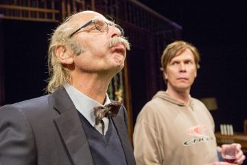 Vanhempi mies, jolla on silmälasit ja rusetti, katsoo ylöspäin huulet supussaan, kun taas nuorempi mies, jolla on rento collegepaita, seisoo hänen takanaan ja tuijottaa huolestuneena eteenpäin.