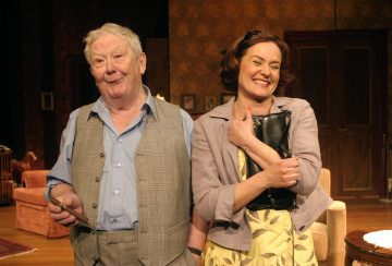 Vanhempi mies harmaassa liivissä ja nainen keltaisessa mekossa ja harmaassa takissa hymyilevät lämpimästi seisoessaan yhdessä viihtyisässä, lämpimästi valaistussa lavastuksessa, jonka taustalla on klassisia huonekaluja.