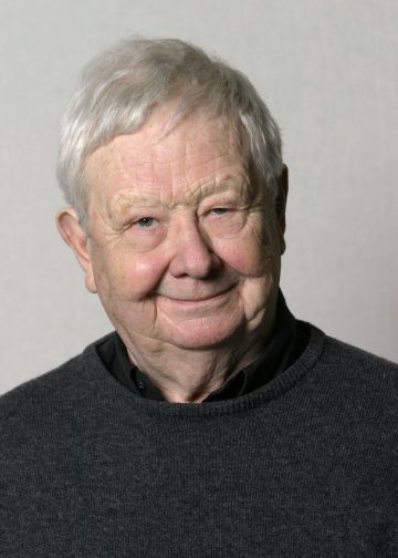 Lyhyet valkoiset hiukset omaava iäkäs mies, jolla on tumma villapaita kauluspaidan päällä, hymyilee lempeästi kameralle vaaleanharmaalla taustalla.