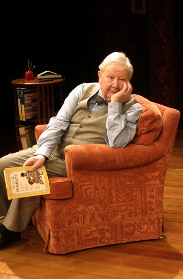 Harmaaliiviin ja siniseen paitaan pukeutunut iäkäs mies istuu oranssissa nojatuolissa, nojaa päänsä käteensä ja näyttää mietteliäältä. Hänellä on kädessään aikakauslehti. Taustalla on sivupöytä, jossa on kirjoja ja punainen omena.