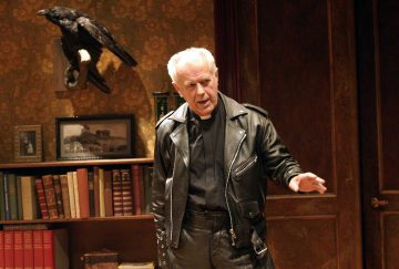 Vanhempi valkotukkainen mies, jolla on musta nahkatakki ja mustat housut, elehtii yhdellä kädellä huoneessa, jossa on kirjahyllyjä, puupaneloidut ovet ja seinälle kiinnitetty korppi.