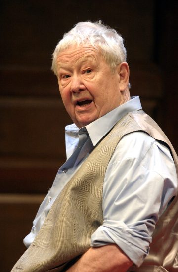 Vanhempi mies, jolla on lyhyet valkoiset hiukset, vaaleansininen paita ja harmaa liivi, katsoo hieman olkansa yli ilmeikkäästi ja suu auki. Tausta on pehmeästi sumea.