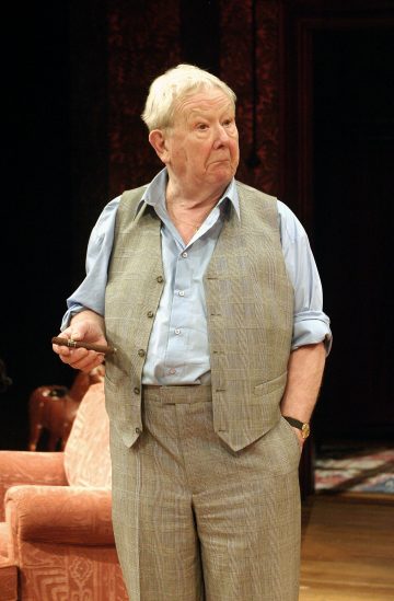 Vanhempi harmaahiuksinen mies, jolla on vaaleansininen paita, harmaa liivi ja harmaat housut, seisoo sisätiloissa pitelemässä esinettä toisessa kädessään ja toinen käsi taskussaan katsellen sivulle.