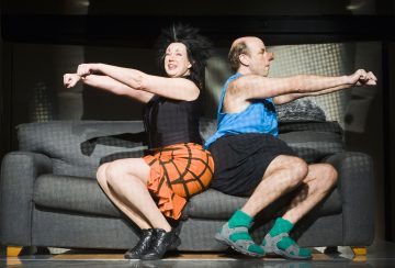 Kaksi ihmistä istuu sohvalla selkä vastakkain ja teeskentelee ajavansa polkupyörällä. Naisella on musta toppi ja oranssi hame, kun taas miehellä on sininen toppi, mustat shortsit ja vihreät sukat sekä sandaalit. Molemmat vaikuttavat energisiltä ja leikkisiltä.