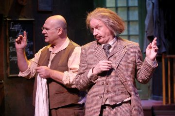 Sisätiloissa seisoo kaksi aikakauden pukuihin pukeutunutta miestä; toisella on kalju, silmälasit ja liivi, ja hänellä on kädessään pyyhe, kun taas toisella, jolla on villi harmaa tukka ja viikset, on ruudullinen puku ja hän elehtii molemmilla käsillään.