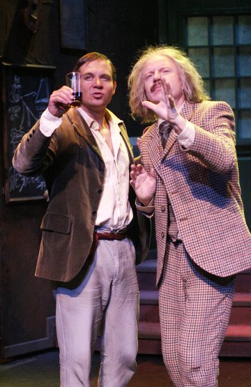 Sisällä seisoo kaksi vanhaan pukuun pukeutunutta miestä. Toinen pitää lasia ylhäällä ikään kuin kohottaisi maljan, ja toinen elehtii kädellään. Molemmat vaikuttavat eloisilta, mahdollisesti näyttelevät näytelmässä, jonka taustalla on liitutaulu ja ikkuna.