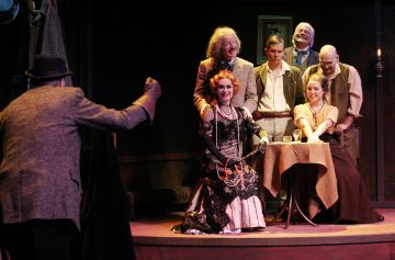 Lavalla esiintyy joukko aikakauden pukuihin pukeutuneita näyttelijöitä; hienostuneeseen pukuun pukeutunut nainen istuu pöydän ääressä toisen naisen kanssa, ja miehet seisovat ja elehtivät vilkkaasti heidän ympärillään dramaattisen valaistuksen keskellä.