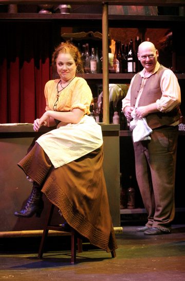 Historiallisiin vaatteisiin pukeutunut nainen istuu baarijakkaralla hymyillen ja kädet ristissä, kun taas baaritiskin takana seisova kalju mies kiillottaa lasia ja hymyilee. Taustalla on hyllyjä, joissa on pulloja ja rekvisiittaa.