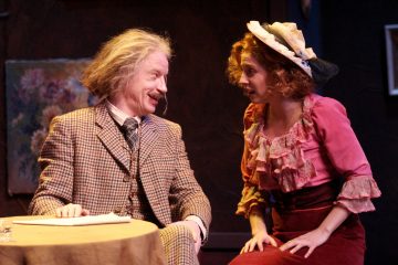 Mies, jolla on villi tukka ja viikset, istuu pienen pyöreän pöydän ääressä ja hymyilee vaaleanpunaiseen röyhelömekkoon ja hattuun pukeutuneelle naiselle, joka kumartuu häntä kohti, ja molemmat keskustelevat vilkkaasti lavalla.