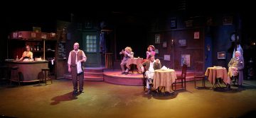 Näytelmäkohtaus kuvaa useita näyttelijöitä hämärästi valaistussa kahvilassa, jossa on pöytiä, tuoleja ja baari. Jotkut näyttelijät istuvat pöydissä, kun taas toiset seisovat tai esiintyvät, mikä luo dynaamisen ilmapiirin.