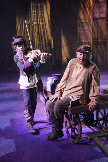 Kaksi näyttelijää näyttämöllä; toinen soittaa viulua korkeaan hattuun ja värikkäisiin vaatteisiin pukeutuneena, toinen istuu puukärryllä talonpoikaisasuun pukeutuneena dramaattisessa valaistuksessa, taustalla maalaismaiset puiset maisemat.