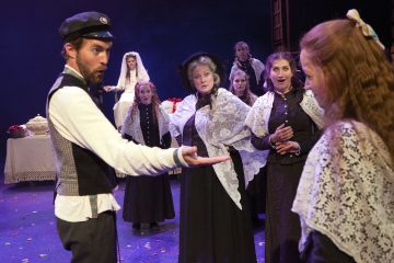 Lakkipäinen mies osoittaa dramaattisella eleellä kohti lavalla olevaa ryhmää vanhanaikaisiin vaatteisiin ja pitsihuiveihin pukeutuneita naisia, joiden taustalla istuu valkoiseen mekkoon pukeutunut nainen. Kohtaus näyttää olevan teatteriesityksestä.