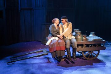 Kaksi aikakauden pukuihin pukeutunutta näyttelijää istuu hymyillen lähekkäin puukärryllä, jonka lavalla on maitotölkkejä, dramaattisen sinisen valaistuksen alla, mikä viittaa maaseutukylään sijoittuvaan teatteriesitykseen.