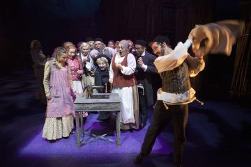 Joukko aikakauden pukuihin pukeutuneita näyttelijöitä kerääntyy pöydän ympärille, jossa on ompelukone näyttämöllä, ja ilmaisee hämmästystä ja jännitystä, kun mies heiluttaa kangasta dramaattisesti. Kohtaus on kirkkaasti valaistu tummaa taustaa vasten.