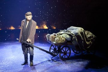 Vanhempi mies kuluneissa vaatteissa ja lippiksessä seisoo lumisella näyttämöllä vetäen suurta puukärryä, joka on täynnä katettuja esineitä, ja taustalla on tumma, savuinen tausta ja putoavia lumihiutaleita.