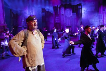 Maalaispukuinen mies seisoo näyttämön etualalla, kädet lanteilla, kun hänen takanaan tanssii joukko miehiä. Taustalla on puurakennuksia ja dramaattinen violetti valaistus.