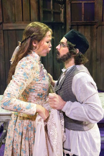 Kukkamekkoon pukeutunut nainen ja vanhanaikaisiin vaatteisiin pukeutunut mies katsovat toisiaan intensiivisesti ja seisovat lähellä toistensa edessä vakavin ilmein, aivan kuin he olisivat kesken kiihkeän keskustelun. Taustalla näkyy maalaismaisia puuseiniä ja ikkunoita.