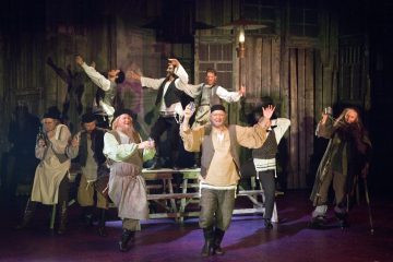 Ryhmä perinteisiin itäeurooppalaisiin pukuihin pukeutuneita näyttelijöitä esiintyy energisesti lavalla, jotkut seisovat pöydillä ja toiset tanssivat iloisin ilmein maalaismaista puista taustaa vasten.