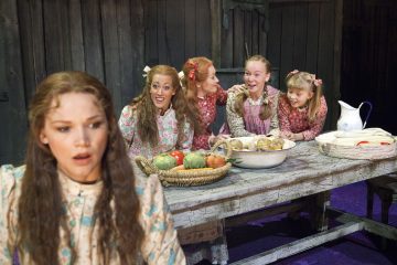 Viisi vanhanaikaisiin mekkoihin pukeutunutta tyttöä istuu maalaismaisen puupöydän ympärillä vihannesten, leivän ja kannun kanssa. Neljä hymyilee ja juttelee pöydän ääressä, kun taas yksi tyttö edessä näyttää huolestuneelta tai järkyttyneeltä.