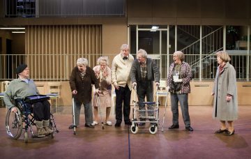 Seitsemän iäkästä ihmistä, joista osa liikkumisen apuvälineiden, kuten pyörätuolin, kävelytuolin ja kainalosauvojen, kanssa, seisovat ja ovat vuorovaikutuksessa tilavassa, hyvin valaistussa sisätilassa, jonka taustalla on moderneja arkkitehtonisia elementtejä.