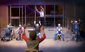 Ryhmä vanhuksia, joista osa on pyörätuolissa, istuu puoliympyrässä kädet kohotettuna, kun edessä oleva henkilö johtaa heitä; toinen henkilö seisoo kaiteen takana ja nostaa myös kätensä ylös.