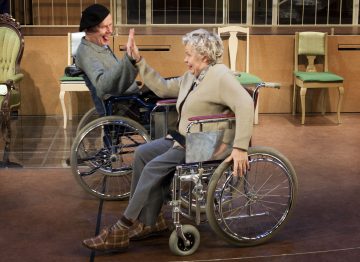 Kaksi pyörätuolissa istuvaa vanhusta hymyilee ja antaa toisilleen peukutuksen tyhjien tuolien ympäröimänä sisätiloissa.