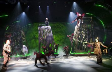 Teatterin näyttämöllä on dramaattinen valaistus, näyttelijät taidokkaissa puvuissa, kaksi suurta häkkimäistä rakennelmaa ja vihreän sävyinen tunnelma, joka viittaa fantasiaan tai maagiseen teemaan. Esiintyjät tanssivat ja ovat tarmokkaasti vuorovaikutuksessa.